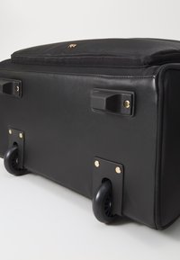 Feketét kerekű bőrönd sima külsejével, aranyszínű fém kiegészítőkkel, masszív oldalsó fogantyúkkal és mintás gumikerékekkel.