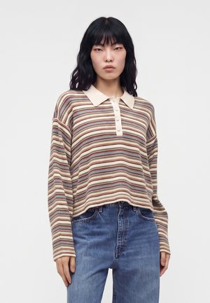Jeune femme aux cheveux noirs porte une chemise beige rayée à manches longues avec col, et un jean bleu taille haute, debout devant un fond uni.