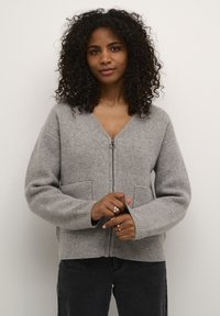 Cardigan lavorato a maglia grigio chiaro con scollo a V e apertura con zip. Dispone di due tasche frontali e polsini a coste. Tessuto morbido, design casual.