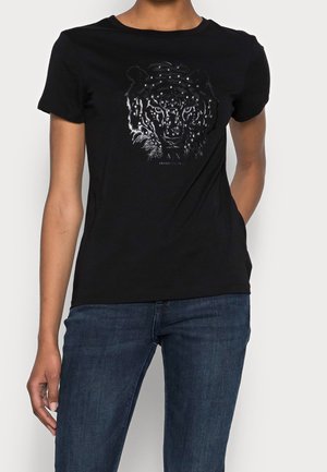 Kobieta ubrana w czarną koszulkę z krótkim rękawem z metaliczną grafiką twarzy lamparta oraz niebieskie dżinsy, stojąca na jednolitym tle.