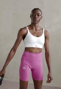 Hvit sports-bh med svarte stropper; rosa høyhalsede shorts; tettsittende design for treningstøy; glatt tekstur. Under Armour-logo synlig.