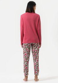 Vrouw die blootsvoets met de rug naar de kijker staat, gekleed in een lange mouwen roze top en een bruine broek met bloemenprint op een witte achtergrond.