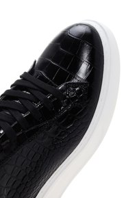 Gros plan d'une basket noire avec une tige en cuir à motif crocodile brillant et des lacets noirs, avec une semelle épaisse en caoutchouc blanc.