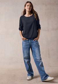 Mørkeblå langærmet bluse med rund halsudskæring, parret med lys blå bredbensjeans og hvide sneakers på en neutral baggrund.
