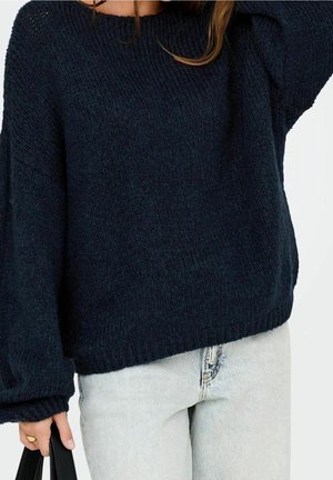 Persoon draagt een losse donkerblauwe gebreide trui en lichtblauwe spijkerbroek, houdt een zwarte handtas in de linkerhand.