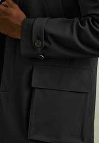 Mustaline väliwear sileda kangaga viimistlusega, millel on patch tasku ja nööpidega mansetid. Disain on sujuv ja minimaalsete aksessuaaridega.