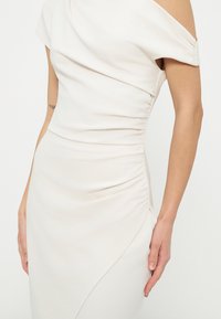 Robe blanche épaules dénudées avec côtés froncés et encolure asymétrique, portée par une personne avec un tatouage visible sur le bras et des ongles neutres.