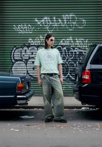 Light green graphic t-shirt with "New Era Cap Co" text, distressed wide-leg jeans, black chunky shoes, and white sunglasses. Urban street setting.