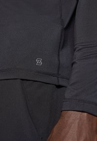 Melna sportiska garroku krekls ar piegulošu dizainu, gludu tekstūru un smalku logotipu apakšējā kreisajā pusē.