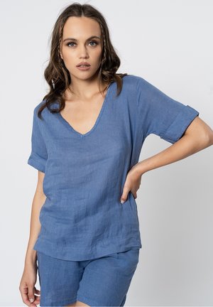 Giovane donna con capelli mossi indossa una maglia azzurra di lino a V larga e pantaloncini abbinati, posando con una mano sui fianchi su sfondo grigio.