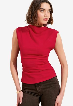 Frau mit welligem braunem Haar, die ein ärmelloses rotes Oberteil und dunkelbraune Hosen trägt, und mit einer Hand seitlich posiert.