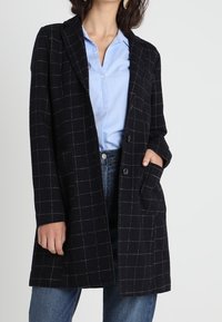 Femme portant un long manteau noir à carreaux par-dessus une chemise bleu clair boutonnée et un jean bleu, avec une main dans la poche du manteau.