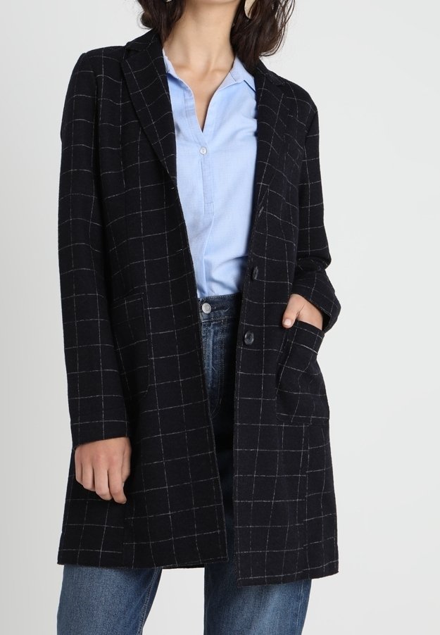 Femme portant un long manteau noir à carreaux par-dessus une chemise bleu clair boutonnée et un jean bleu, avec une main dans la poche du manteau.