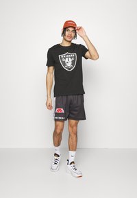 Camiseta negra con el logo blanco de "RAIDERS", combinada con pantalones cortos deportivos grises que presentan detalles en rojo y blanco. Los accesorios incluyen una gorra naranja.
