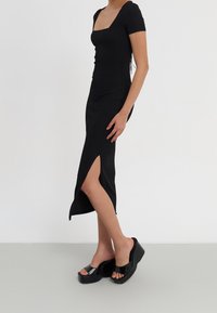Smal passform svart midi-klänning med korta ärmar och sidoslits, bärs med svarta plattformssandaler med öppen tå mot en neutral bakgrund.