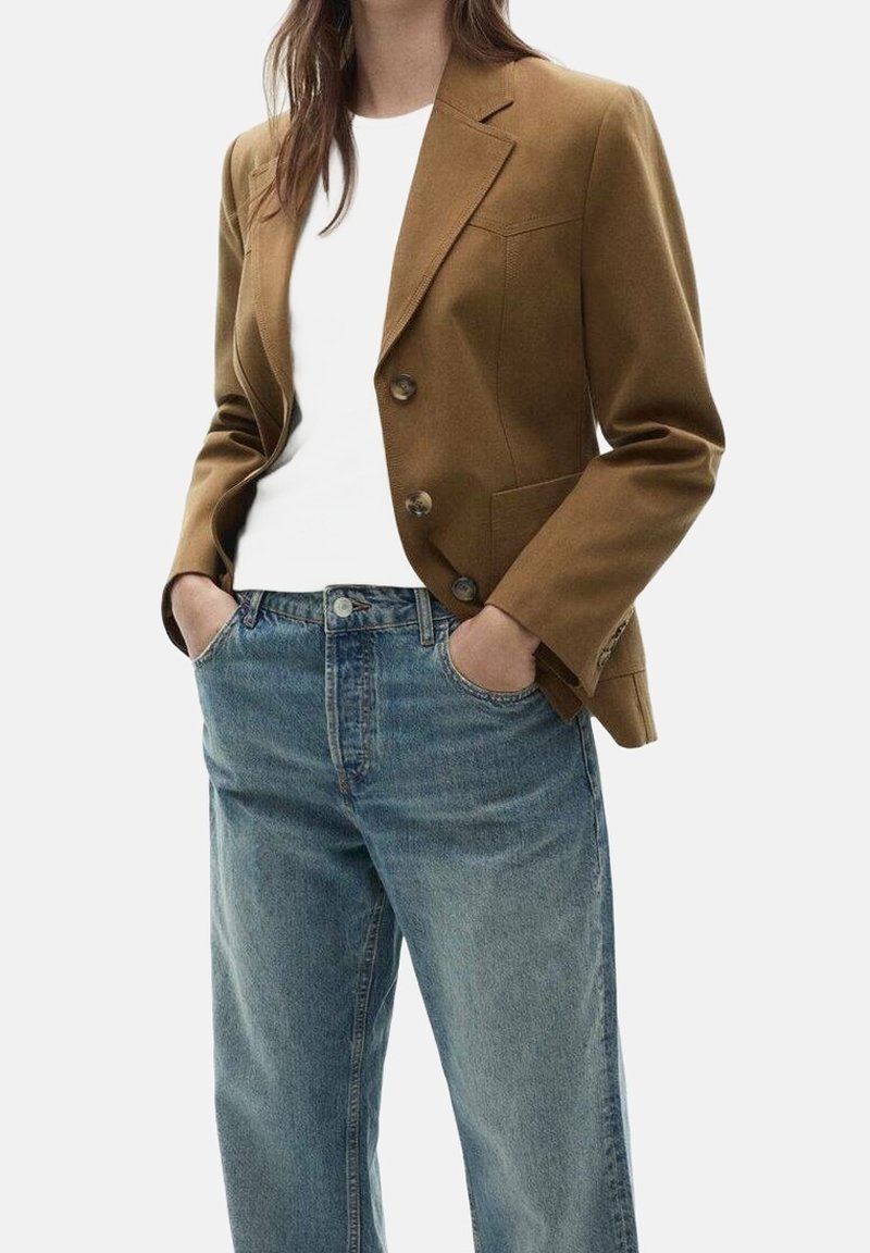 Femme portant un blazer marron boutonné sur une chemise blanche, les mains dans les poches d'un jean bleu ample.