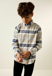 Camisa de manga larga a cuadros en beige con rayas azules, grises y blancas. Presenta un cuello abotonado y cierre de botones con un ajuste relajado.