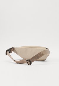 Beige szövet deréktáska ívelt formával, cipzáros záródással és állítható pánttal, amelyen dombornyomott logó található.
