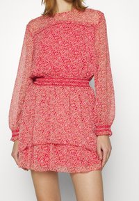 Mini robe fleurie rouge avec manches longues et transparentes, taille smockée et volants en couches. Caractérisée par un tissu délicat à motifs et une texture légère.