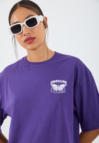 Camiseta de manga corta morada con gráfico de mariposa blanca y texto en el pecho. El modelo lleva gafas de sol cuadradas blancas. Textura de algodón suave.