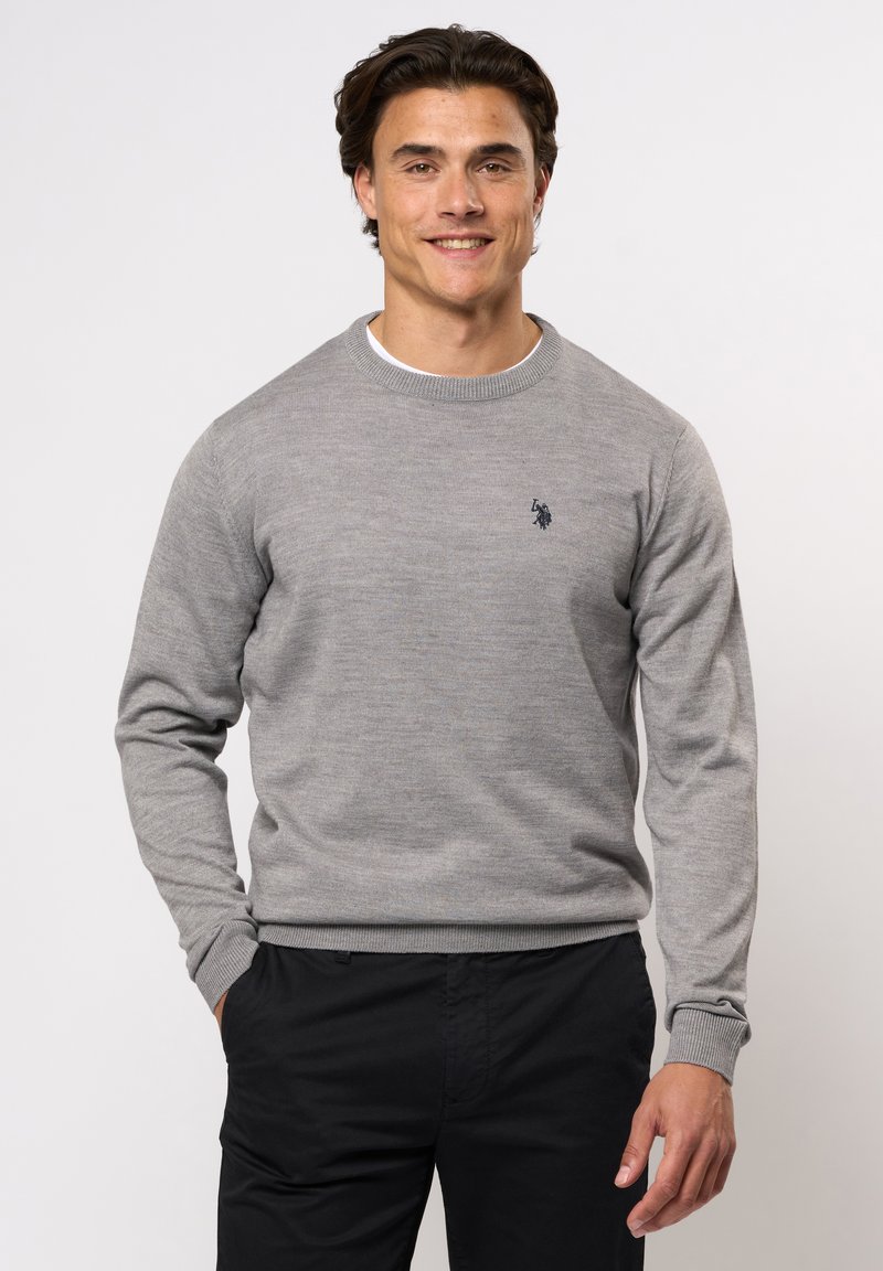 Grijze gebreide trui met een ronde hals, geribde manchetten en zoom, voorzien van een klein marineblauw logo op de linkerborst. Gedragen over een wit shirt.