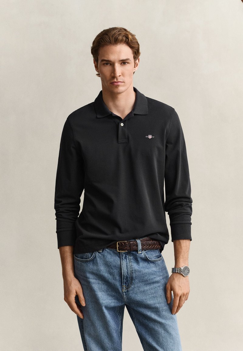 Joven con cabello castaño que lleva una camiseta polo negra de manga larga, vaqueros azules, cinturón marrón y reloj de pulsera plateado sobre un fondo liso.