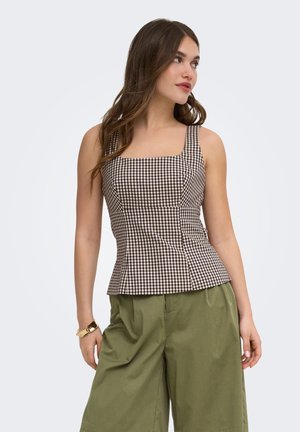 Femeie purtând un top fără mâneci în carouri maro și alb și pantaloni largi verde măsliniu, uitându-se spre dreapta, cu o brățară aurie la încheietură.