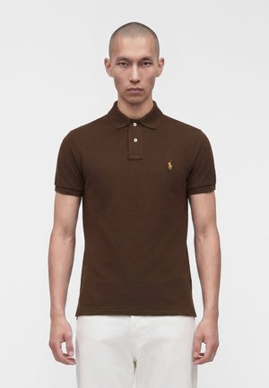 Homme au crâne rasé portant un polo marron à manches courtes avec un logo sur la poitrine et un pantalon blanc, debout sur un fond blanc uni.