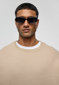 Zwarte ovale zonnebril, beige oversized sweatshirt met een ronde halslijn, gedragen over een wit t-shirt. Glad textuur en minimale detaillering.