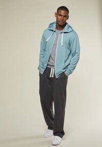 Mann steht mit den Händen in den Taschen, trägt einen hellblauen Reißverschluss-Hoodie, ein graues T-Shirt, dunkle graue Jogginghosen und weiße Sneakers auf beigem Hintergrund.