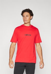 Raudona trumpų rankovių marškinėlių suapvalinta kaklo iš 100% medvilnės, su juodu "adidas" logotipu, esant centru po kaklu.