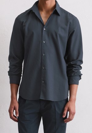 Man die een donkergrijs, lange mouwen, knoop overhemd met open kraag en bijpassende donkere broek draagt tegen een effen lichte achtergrond.