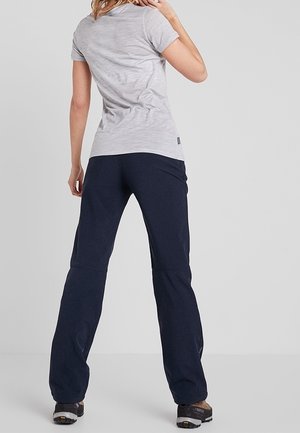 Femme portant un t-shirt gris clair à manches courtes, un pantalon bleu marine et des chaussures de randonnée, debout de dos face à la caméra sur un fond uni.