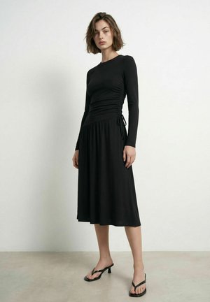 Femme portant une robe midi noire à manches longues avec des fronces sur les côtés et des sandales noires à talons, debout sur un sol uni devant un mur blanc.