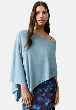 Tânără cu păr lung și castaniu, purtând un poncho asimetric bleu și o fustă închisă cu modele florale albastre.