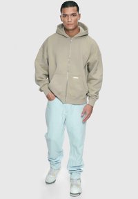Khaki hettejakke med glidelås i løs passform, stor hette og frontlommer, sammen med lyseblå jeans og hvite sneakers med gule detaljer.