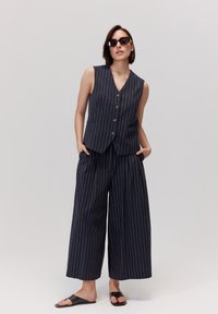 Granatowa kamizelka w prążki z guzikami, zestawiona z szerokimi spodniami, wykonanymi z gładkiego materiału. Model nosi okulary przeciwsłoneczne i czarne sandały.