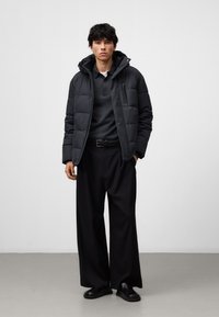 Giacca nera imbottita con cappuccio, dotata di chiusura con zip frontale e tasche laterali, indossata sopra un maglione grigio e pantaloni neri a gamba larga.