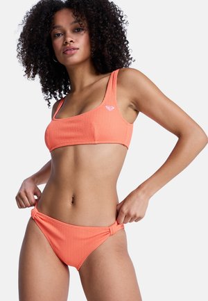 Frau, die korallrote Bikinihosen mit Textur anpasst, trägt passendes Oberteil mit kleinem Logo auf der linken Brust, vor weißem Hintergrund.