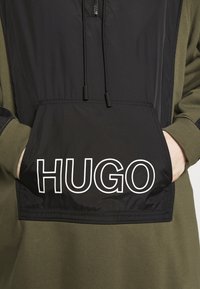 Huppari tummanoliivinvihreänä ja mustana, jossa etutasku valkoisella "HUGO"-logolla, vetoketjukiinnitys ja säädettävät nyörit.
