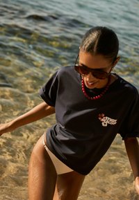 Mørkeblå bomulds T-shirt med blomster grafik og teksten "STUDY IN BLOOM", parret med en cremefarvet bikini bund. Båret med en rød perlekæde.
