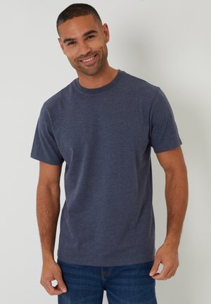 Smilende mand med kort hår iført en ensfarvet mørkeblå kortærmet t-shirt og blå jeans, stående mod en hvid baggrund.