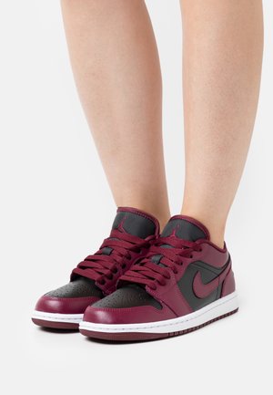 Personne portant des baskets Nike basses noires et rouge foncé avec des lacets, debout sur une surface blanche.