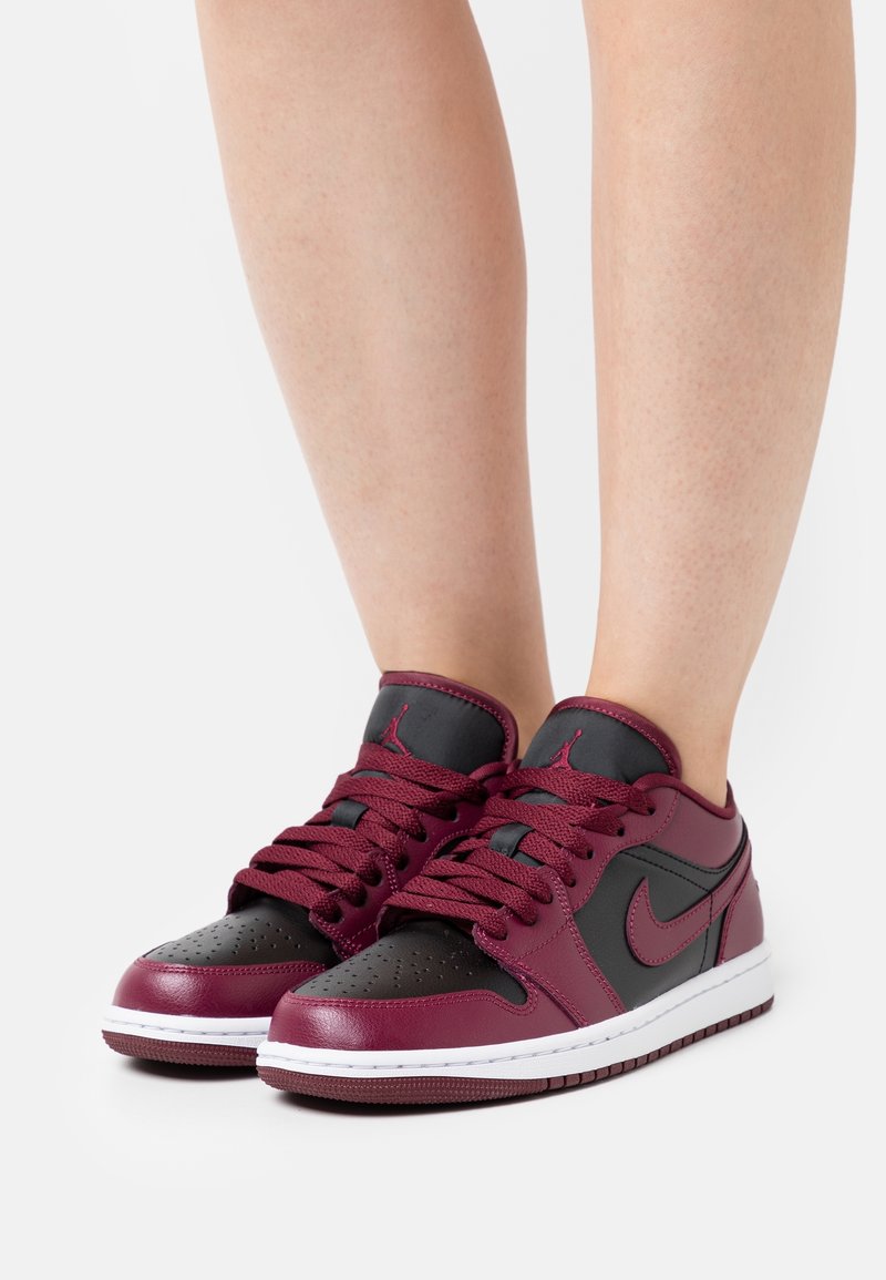 Personne portant des baskets Nike basses noires et rouge foncé avec des lacets, debout sur une surface blanche.