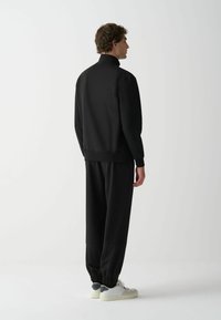 Fekete pulóver magas gallérral, laza fazonban, bordázott mandzsettákkal, párosítva azonos fekete melegítőnadrággal és fehér sportcipőkkel, szürke részletekkel.