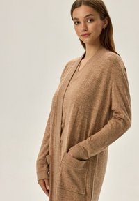Beżowy długi kardigan wykonany z teksturowanej dzianiny, z dekoltem w kształcie V, otwartym przodem i dwiema przednimi kieszeniami naszytymi. Miękki i luźny fason.
