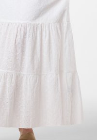 Abito midi bianco in cotone con design ad occhiello, strati a balze e una texture morbida. L'orlo si trova appena sopra la caviglia.