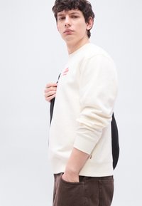 Νεαρός άνδρας που φοράει κρεμ φούτερ και καφέ παντελόνι, κρατώντας μαύρο σακίδιο στον ώμο, κοιτάζοντας προς την κάμερα.