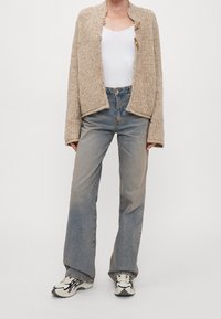 Beige stickad cardigan med avslappnad passform, kombinerad med ljusblå jeans och flerfärgade sneakers. Anmärkningsvärda träknappdetaljer.