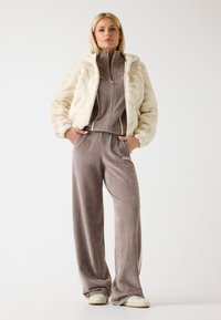 Giacca in pelliccia ecologica crema con un design testurizzato, abbinata a una maglia zip in velluto taupe e pantaloni larghi coordinati, con dettagli del logo.